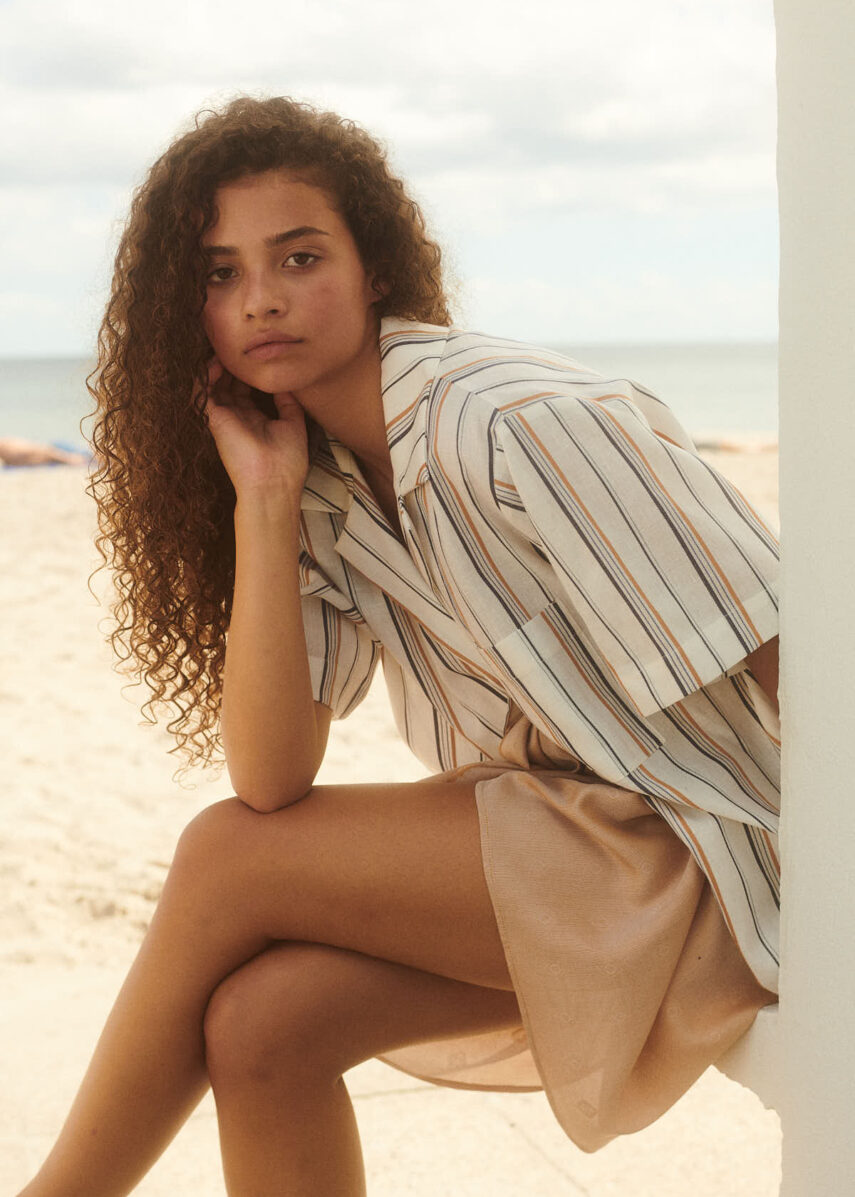 Girl sitting at the beach wearing Alzada Shirt from WAY. A striped beach shirt in linen. She wears a golden sarong around her waist.