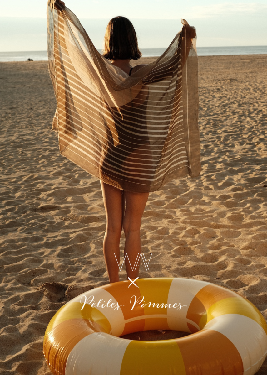 Girl on the beach holding a brown sarong in front of a yellow pool float from petites pommes